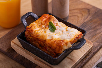 A hearty serving of classic lasagna presented in a black ceramic dish, garnished with a fresh basil leaf. The dish is placed on a wooden board with a glass of orange juice and salt and pepper shakers 