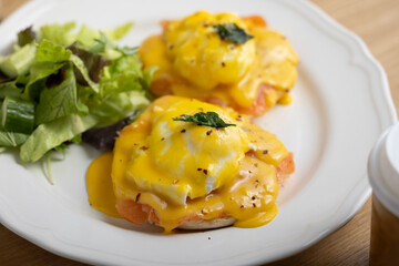 A delicious plate of eggs Benedict topped with rich hollandaise sauce, accompanied by a fresh side salad. The dish is beautifully presented on a white plate, placed on a vibrant outdoor table setting.