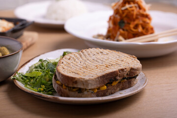 A wholesome sandwich filled with spinach, corn, and cheese, served on whole grain bread with a side of fresh green salad. The sandwich is placed on a rustic white plate with salt and pepper shakers