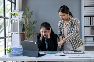 Two women in business attire are sitting at a desk with a laptop and papers