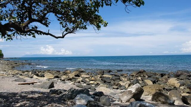 A sunny day on a rocky beach with calm waves against a backgrounds of blue sky and sea in Manatuto, Timor Leste.
