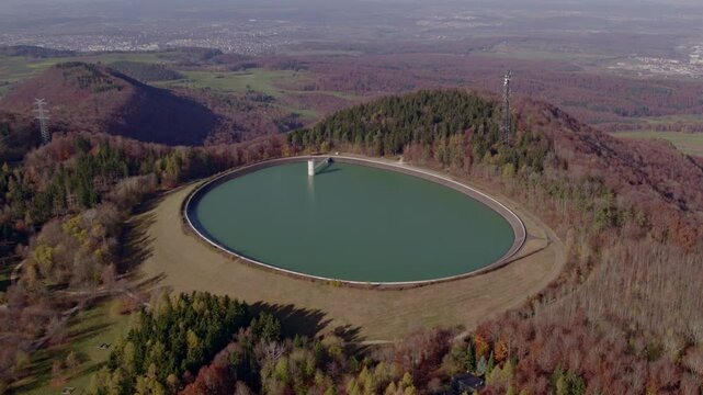 Aerial view of water storage basin surrounded by forest, hill, and mountain in autumn, Eningen unter Achalm, Baden-Wurttemberg, Germany.