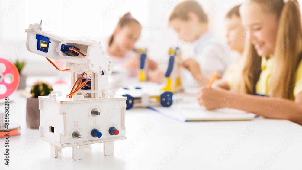 Poster A close-up shot of a white robotic arm on a table in a classroom setting. The robot is in focus and there are blurred children in the background who are building or working on projects.