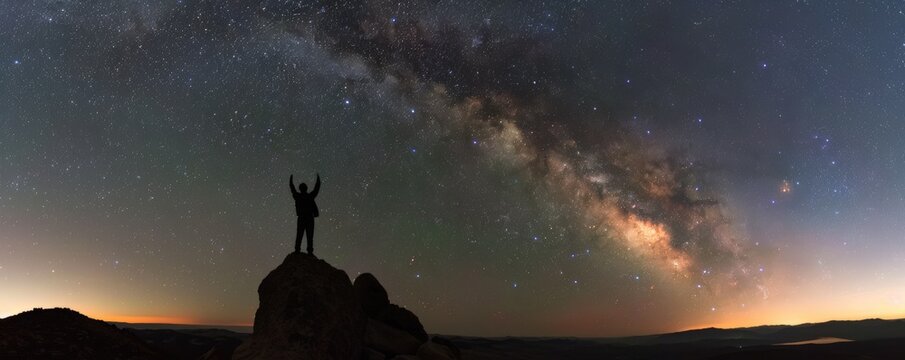 Silhouette of a person on a rock under a stunning Milky Way sky at twilight, capturing the beauty of the stars and universe.