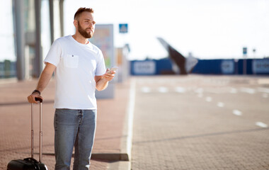 Male tourist arriving at airport, ordering taxi by phone and standing with baggage, free space