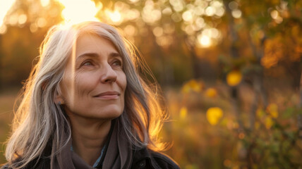 A woman with gray hair smiles softly as she gazes upwards in a sunlit park, her face illuminated by the gentle golden light of the setting sun.