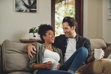 Happy, interracial couple and drinking coffee in home, talking or bonding together for love in living room. Tea cup, man and funny woman in conversation for joke, story or laughing at comedy on sofa