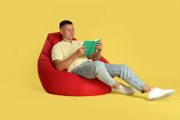 Handsome man reading book on red bean bag chair against yellow background