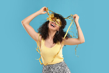Happy young woman wearing carnival mask and tinsel on light blue background