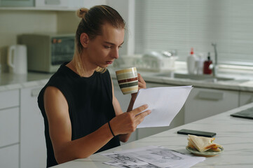 Young model examining sketches while drinking coffee in the kitchen