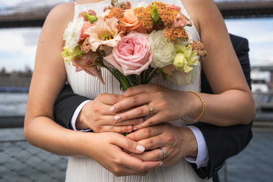 A groom and a bride embrace each other holding hands while simultaneously holding a bouquet of flowers on their wedding day.