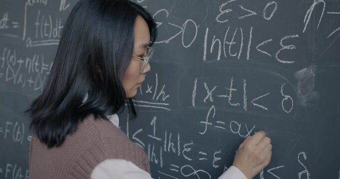 Pensive Asian woman mathematician writing formula on chalkboard thinking focused on education and science development. Youth and knowledge concept.