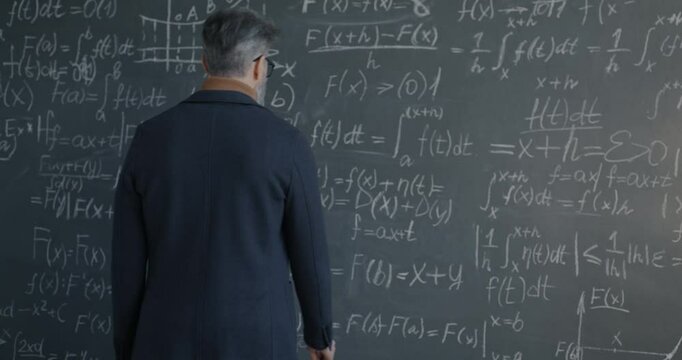 Back view of man professor walking to chalkboard thinking and underlining formula focused on maths theory development. Science and education concept.