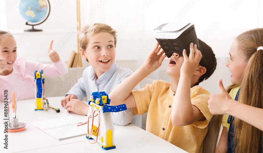 Fototapete A group of children in a classroom are working on a project. One of the children is wearing a virtual reality headset while the others look on. They are all smiling and having fun.
