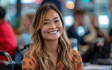 A smiling woman with long brown hair sits in a wheelchair in a brightly lit office setting. She is wearing a floral shirt and surrounded by colleagues in a diverse workplace