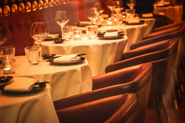 Served tables with white tablecloths with cutlery, plates, glasses, napkins inside a luxury rich restaurant in the evening. Empty seats on a table. 