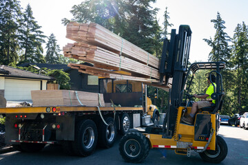 Forklift unloading a load of lumber from a flatbed delivery truck in a residential neighborhood,...
