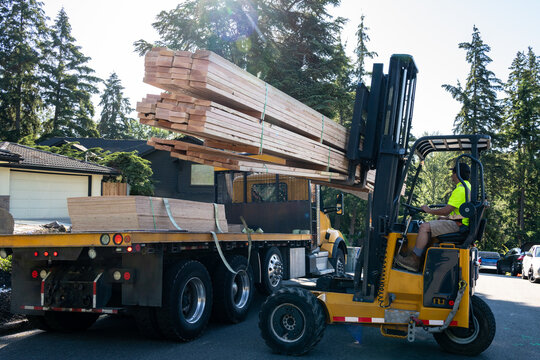 Forklift unloading a load of lumber from a flatbed delivery truck in a residential neighborhood, sunny summer construction season

