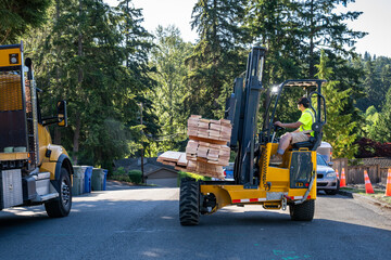 Forklift unloading a load of lumber from a flatbed delivery truck in a residential neighborhood, sunny summer construction season
