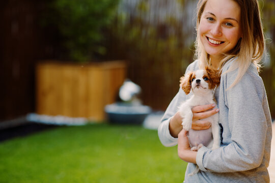 A happy millennial woman smiles holding her adorable Blenheim Cavalier King Charles Spaniel puppy in her green backyard, celebrating love, friendship, bonding, joy of on summer Gotcha day.