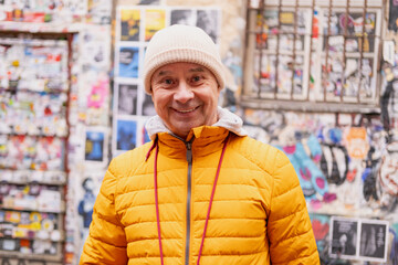 Cheerful middle-aged man, 65 in winter bright yellow puffer jacket and beanie, smiling on urban...
