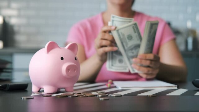 Seated at the kitchen table, a young woman counts banknotes. Piggy bank and scattered coins on the foreground, selective focus. Budgeting and savings concept