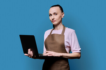 Portrait of 30s woman in apron holding laptop on blue background