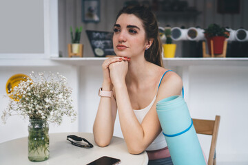 young woman in stylish activewear sits pensively at outdoor café, her yoga mat resting nearby. She...