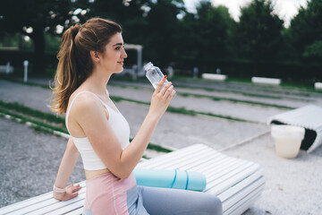 young woman takes break from her exercise routine, sitting on park bench with a yoga mat and a bottle of water. She smiles contentedly, epitomizing healthy living and the joy of outdoor activities.