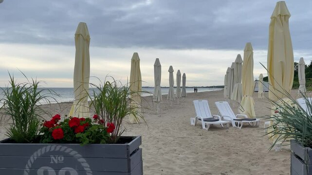 Sunbeds and beach umbrellas on sandy beach. Distant view of a couple walking towards the horizon. Calm, idyllic, resort vibes. Summer vacation concept for tourism. 4K Video.