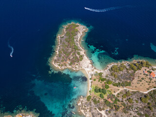 Aerial view of an island on Sithonia Peninsula, Greece, with beaches and rocks, surrounded by crystal-clear waters. Yachts and boats anchor in turquoise waters. Nearby islets and reefs visible