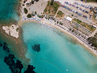 Aerial view of a vibrant beach in Sithonia, Greece, with colorful umbrellas, sunbeds, and beachgoers enjoying the clear turquoise waters, capturing the lively summer atmosphere