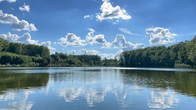 Time-lapse of white clouds moving over peaceful, calm waters of a forest lake. Summer landscape with blue sky and vivid green trees up to the horizon. Concept of calmness, relaxation or mindfulness.