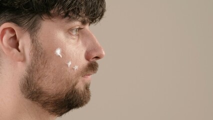 Obraz premium Young adult man applies aftershave cream moisturizer looks at camera, isolated on white background. Side view.