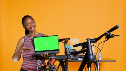 African american woman holds copy space isolated screen laptop, using it to advertise bike repair shop, studio background. Mechanic promoting bicycle fixing service with chroma key notebook
