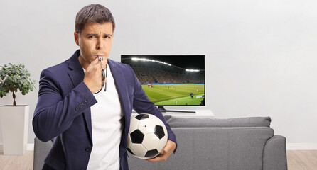 Casual young man holding a football and blowing a whistle in front of tv