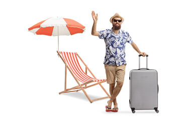 Full length portrait of a bearded man with a suitcase next to a beach chair waving at camera