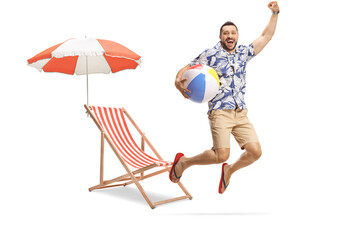 Young excited male tourist holding a ball and jumping in front of a beach chair