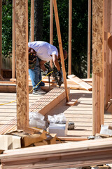 Fototapeta premium Construction worker in toolbelt and hard hat working with a pneumatic nail gun on new house construction wood framing, summer construction season 