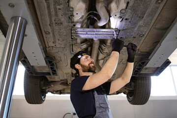 Professional mechanic working underneath car on lift in auto repair shop wearing gloves and headlamp smiling.Focused repairman performing maintenance, ensuring vehicle safety and functionality. © sofiko14