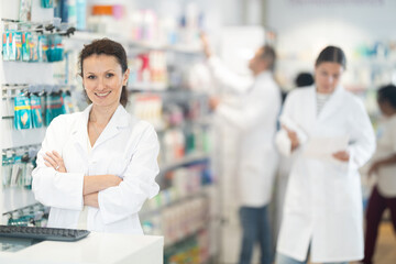 Positive middle-aged female pharmacist standing at the desk using keyboard in chemist's shop