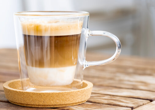 Cup of coffee with milk and froth in transparent glass on the wooden table.