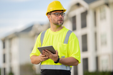 A man in a yellow safety vest is looking at a tablet while wearing a hard hat. He is focused on the...