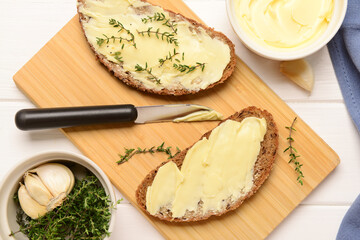Board of bread slices with fresh butter and garlic on white wooden background
