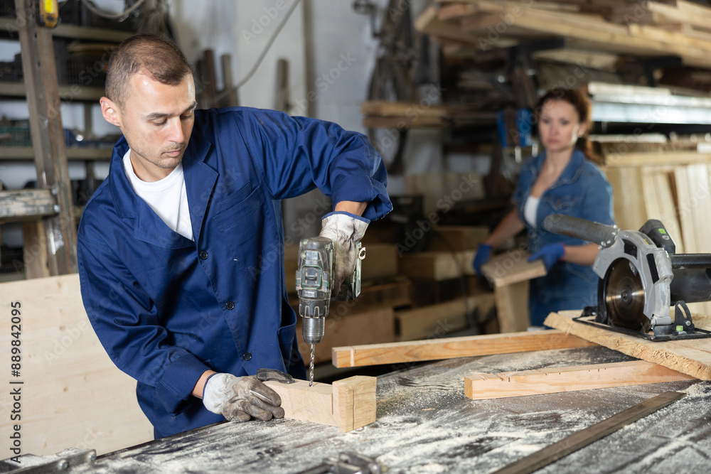 Wall mural male employee of carpentry workshop uses drill to make holes in wooden billet, drill surface, prepar