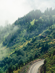 Fototapeta premium Winding mountain road through lush green forest partially covered in mist. Scenic view of curving highway amidst dense foliage, creating a serene and mysterious atmosphere. Perfect for travel and