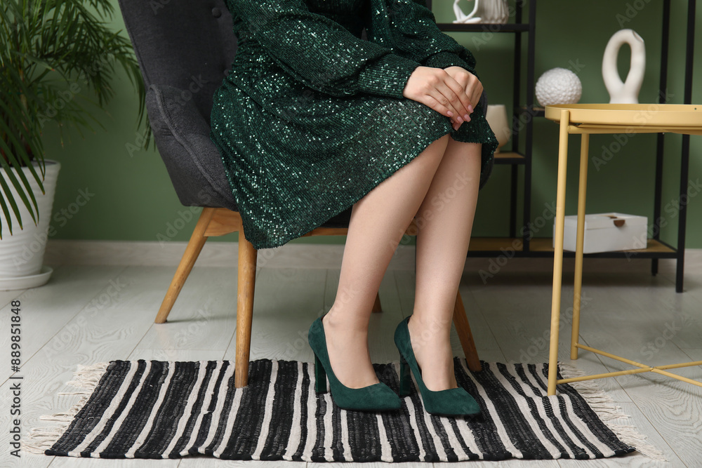 Wall mural Young woman sitting in armchair and trying on green high heeled shoes at home