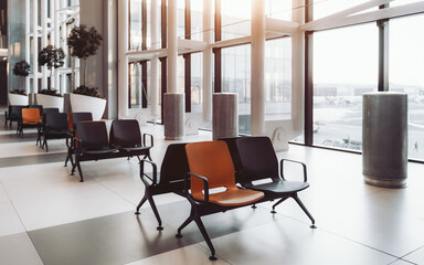 A modern airport waiting area with rows of black and orange seats; potted plants and metal trash cans. Large windows allow natural light to flood the space, showcasing the view of the runway outside