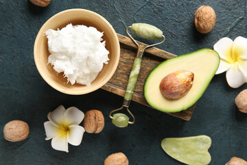 Bowl of shea butter with facial massage tools, avocado, plumeria flowers and nuts on dark background