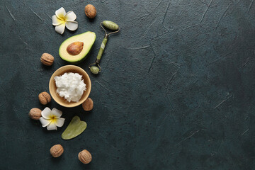 Bowl of shea butter with facial massage tools, avocado, plumeria flowers and nuts on dark background